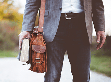 A man is walking with a messenger bag and a book in his hand.